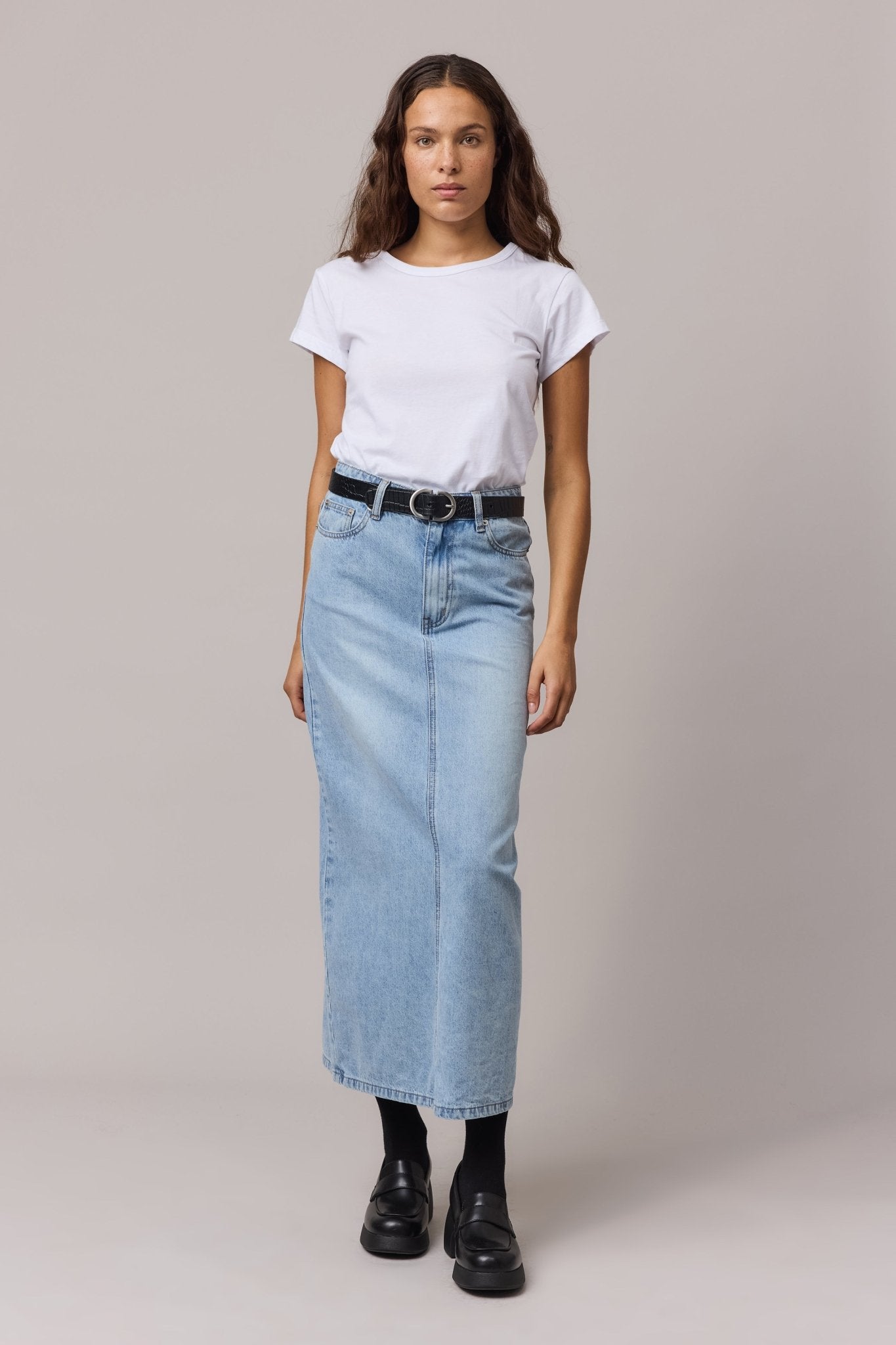 A woman wearing a white organic cotton Girlfriend Tee with a classic crew neck, styled with a light-wash denim maxi skirt and black boots.