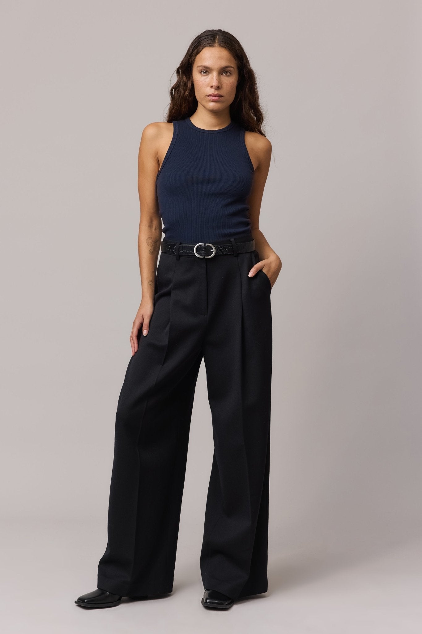 Full-body view of the woman in the indigo tank top and wide-leg black trousers, styled with a black belt.