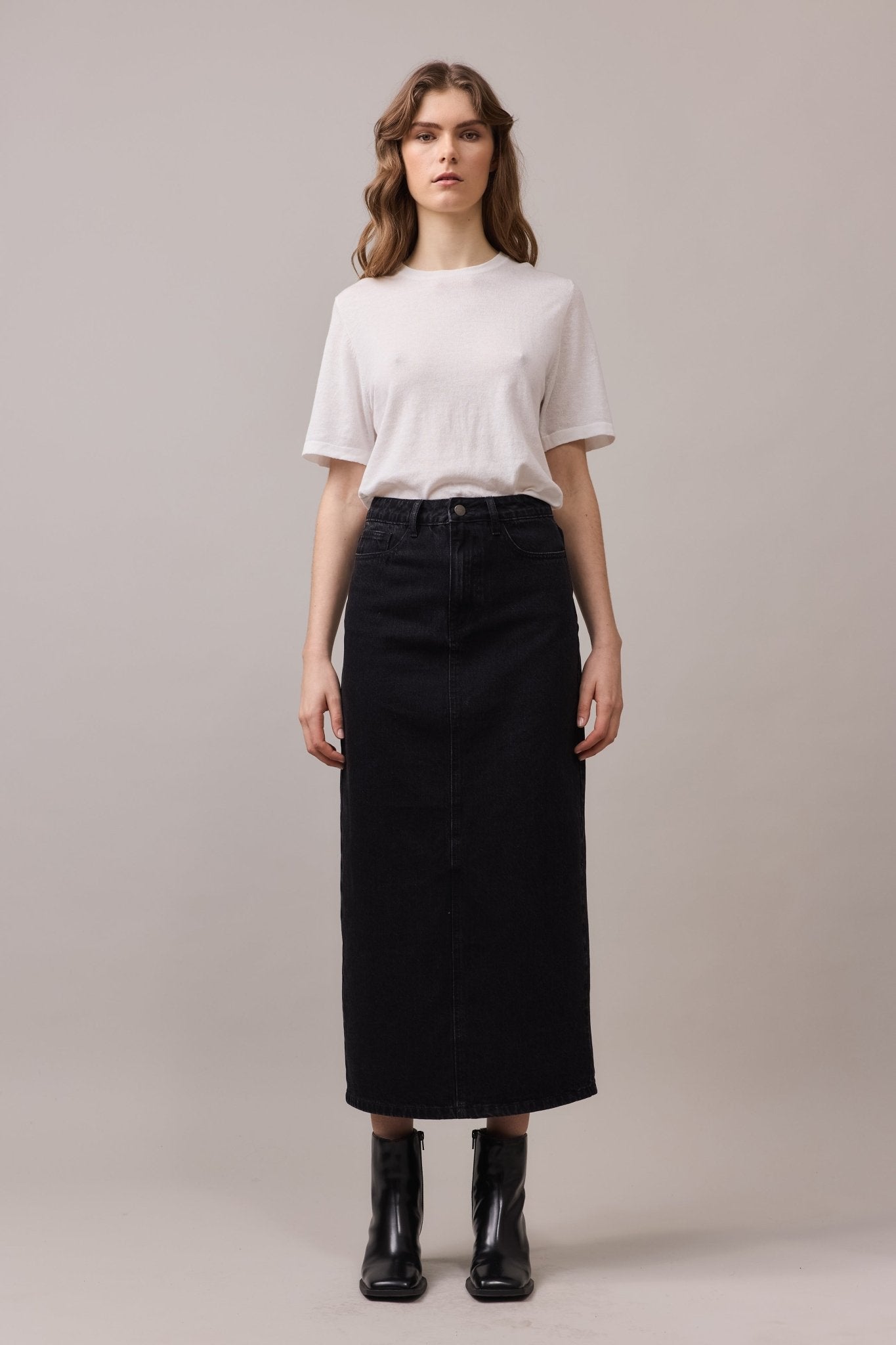 Front view of the woman in a white shirt and black boots, allowing a clear look at the maxi denim skirt’s five-pocket styling and clean lines.