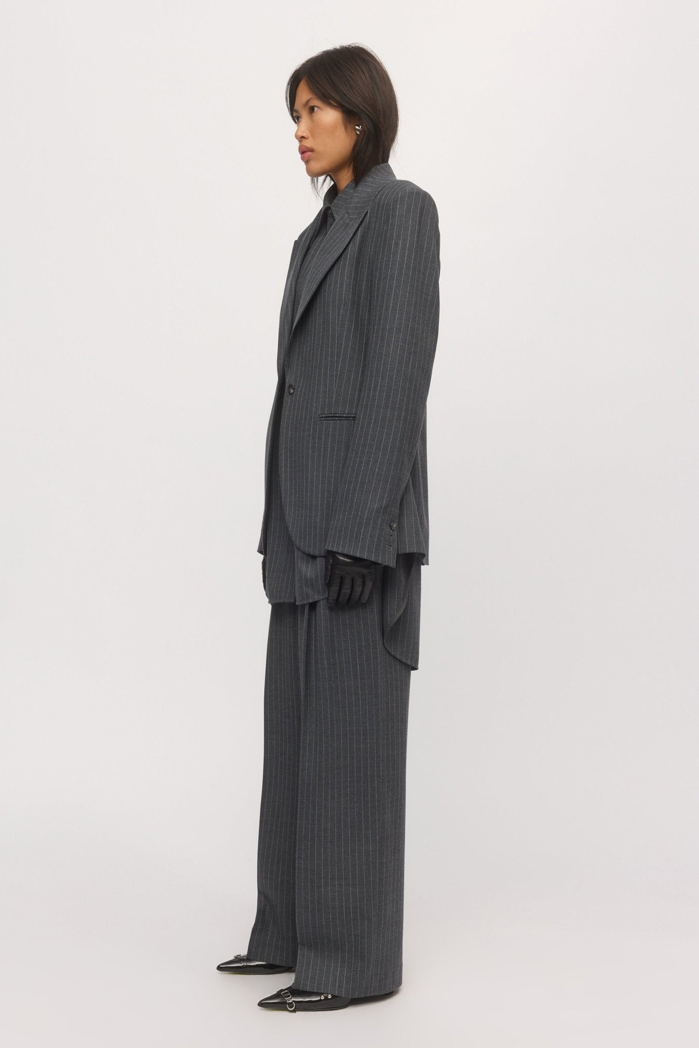 Side view of a woman in a graphite striped blazer, showcasing the blazer’s sharp tailoring, curved hem, and relaxed drape over matching trousers.