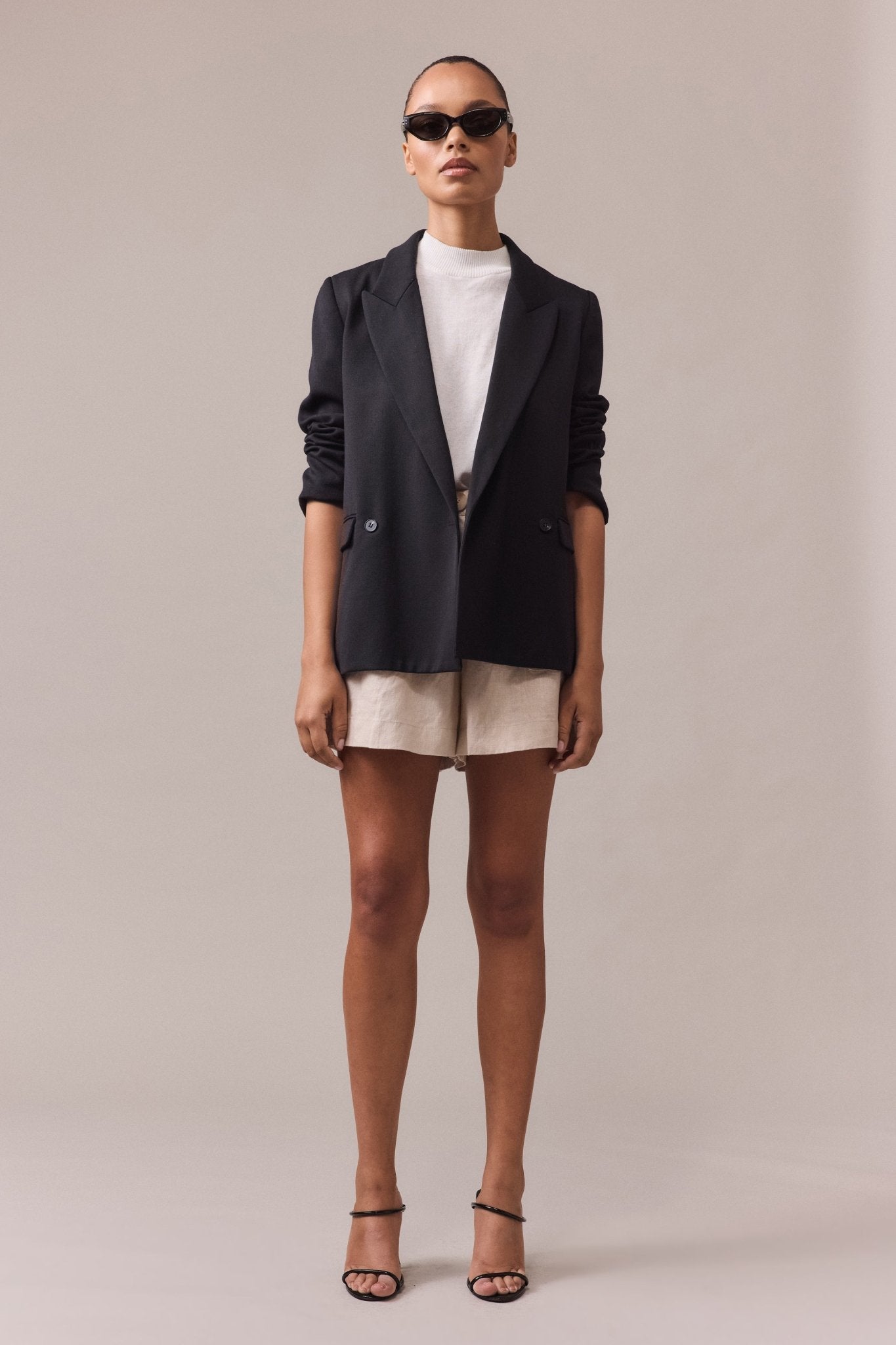 Woman wearing sand Soma shorts and a white top, layered with a black blazer and sunglasses for a chic look.