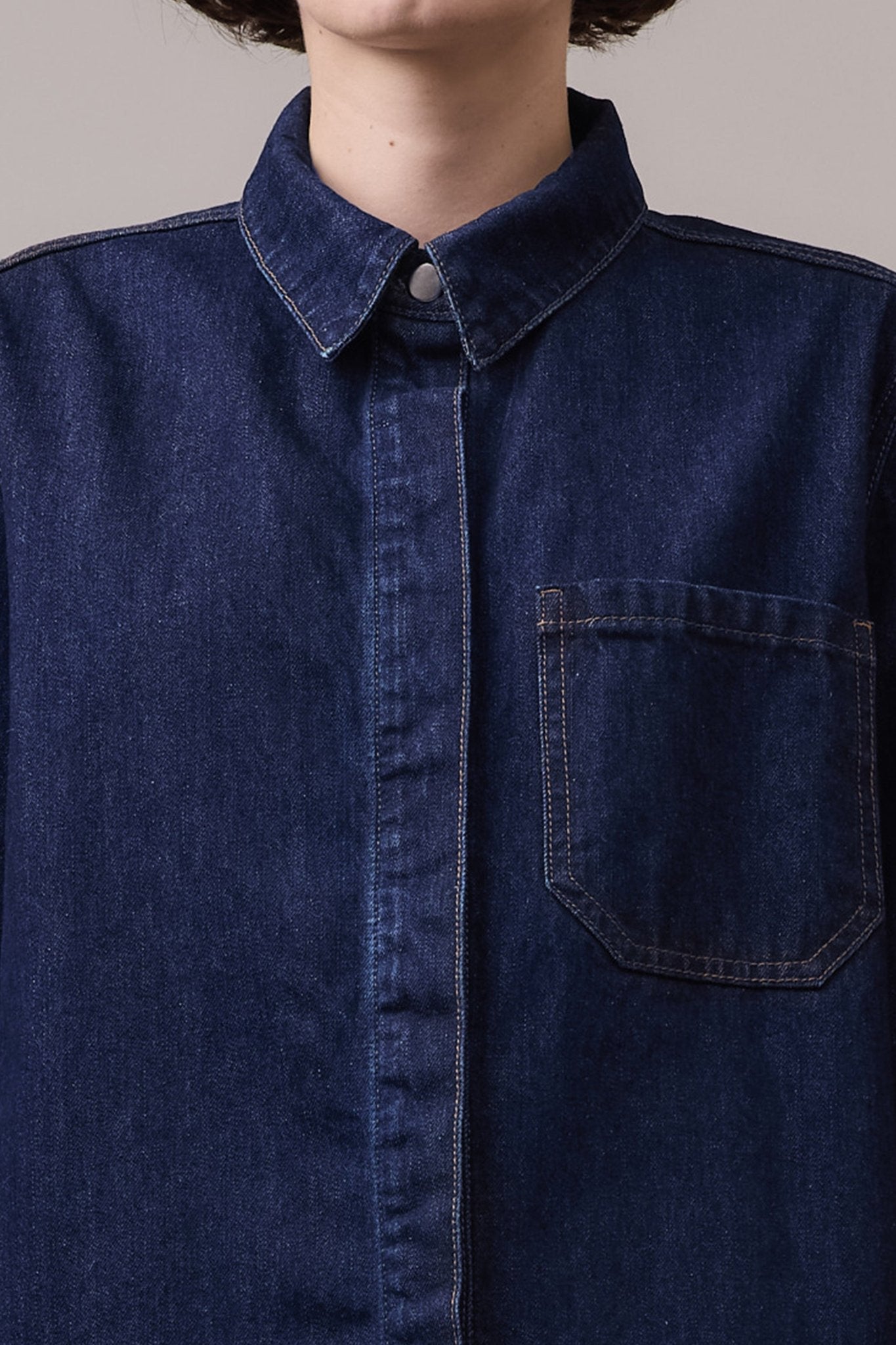 A close-up of a dark denim shirt featuring a classic pointed collar, a front button placket, and a single chest patch pocket with visible contrast stitching.