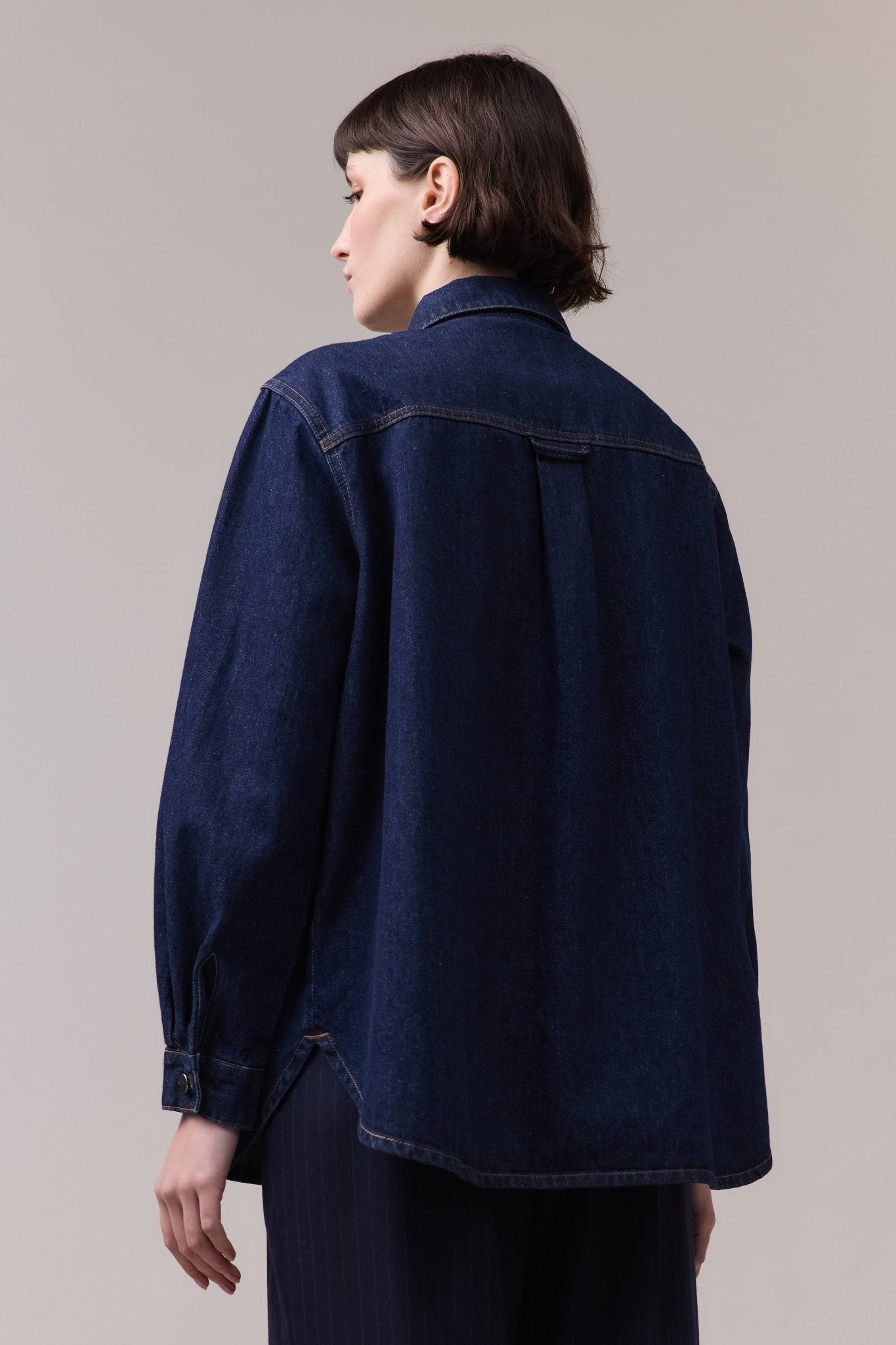 Woman wearing a dark denim shirt with a relaxed fit. The shirt drapes loosely, with a clean back panel and subtle stitching details, creating a casual yet refined look